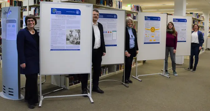 Das Team des Projekts ?GENIAL forschen sowie Bibliotheksdirektor Fabian Franke stehen neben Plakaten bei der Er?ffnung der Posterausstellung.