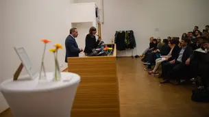 The photo shows David Frum being introduced by Prof. Dr. Christine Gerhardt. Both are standing behind the lectern. The first few rows of the audience are also visible.