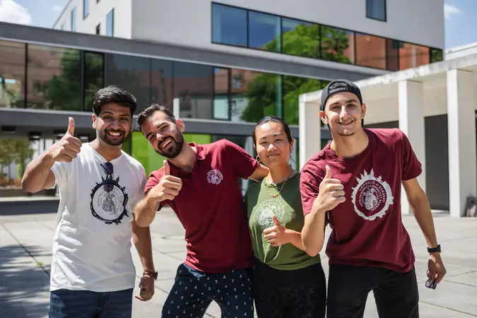 Studierende zeigen vor einem Universit?tsgeb?ude den Daumen nach oben.